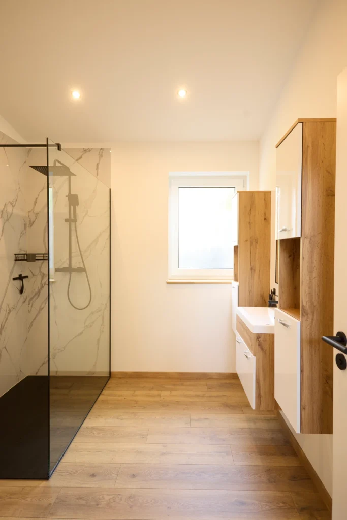 Badezimmer mit Dusche links, Fenster und Waschtisch rechts, Boden in Holzoptik