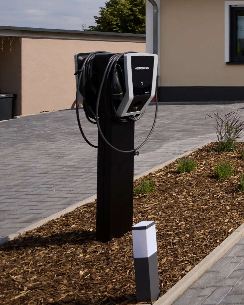 E-Ladesäule mit Ladekabel auf einem Pfosten im Beet neben der gepflasterten Einfahrt