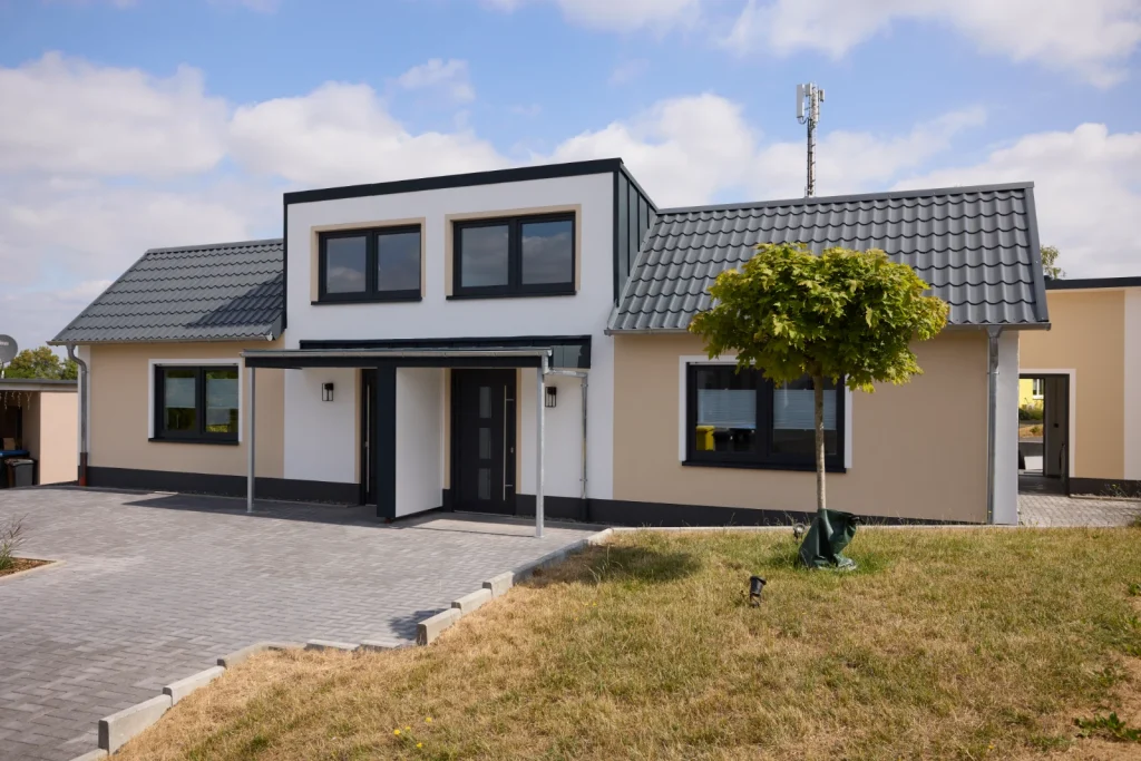 Neu gebaute Doppelhaushälfte mit beigefarbener Fassade, anthrazitfarbenem Dach, Vordach über den Eingängen und kleinem Baum im Vorgarten