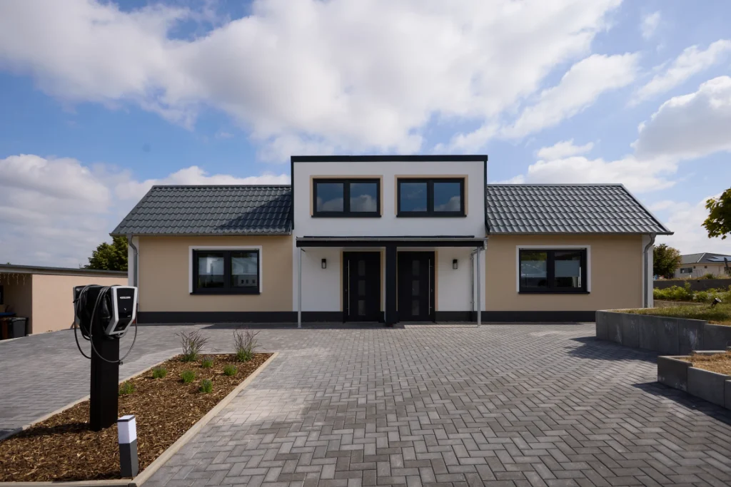 Vorderansicht des Hauses mit zwei Eingangstüren unter einem Vordach, gepflasterter Einfahrt und E-Ladesäule