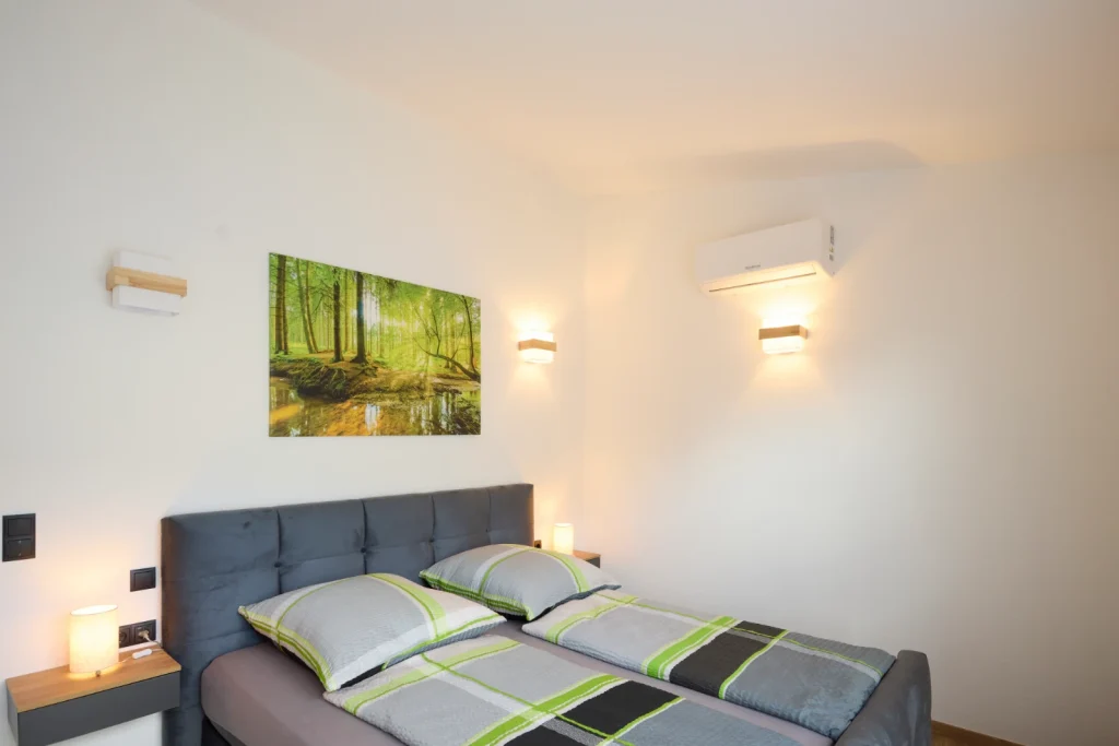 Schlafzimmer mit grauem Polster-Doppelbett, Waldbild an der Wand, warmen Wandleuchten und Klimaanlage