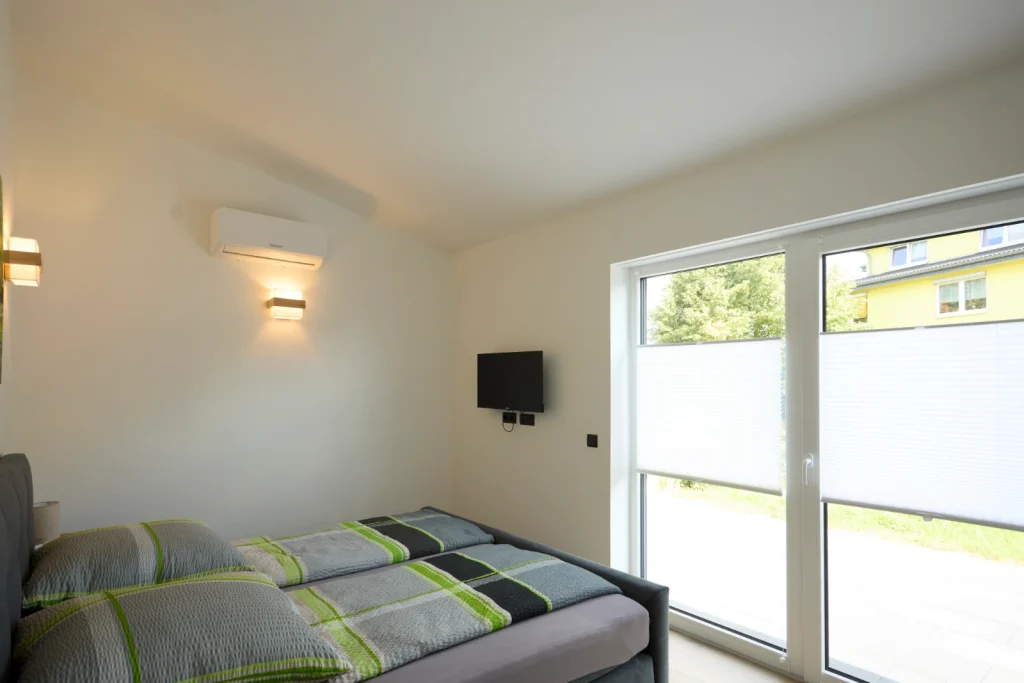 Schlafzimmer mit Doppelbett, Fernseher an der Wand und großer Glastür zur Terrasse