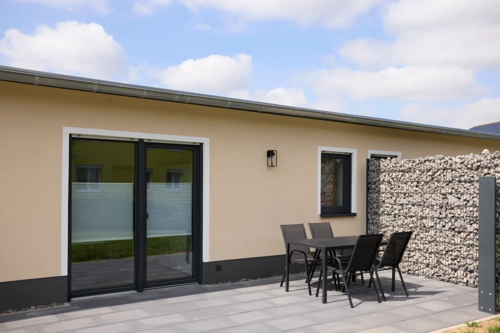 Terrasse mit Tisch und vier Stühlen vor der großen Glastür, seitlich eine Gabionenwand als Sichtschutz