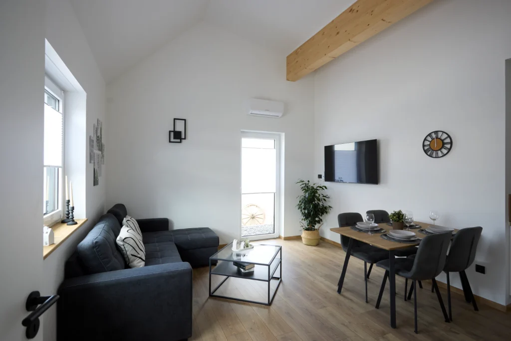 Helles Wohnzimmer mit dunkelgrauem Ecksofa, Couchtisch, Fernseher und gedecktem Esstisch unter einer hohen Decke mit Holzbalken